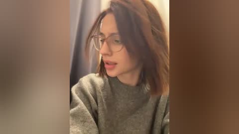 Media: Video of a young woman with medium-length brown hair, wearing glasses, and a grey sweater, looking down, in a blurry, dimly lit room with brown and beige tones.