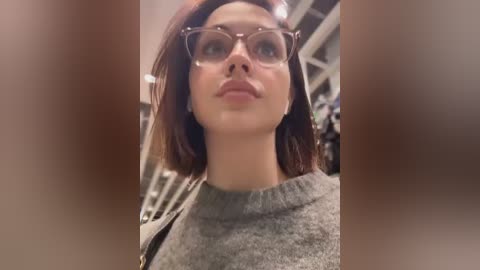 Media: Video of a young woman with light skin and straight brown hair, wearing large, clear-framed glasses and a gray sweater. She stands indoors with blurred background suggesting a retail store.