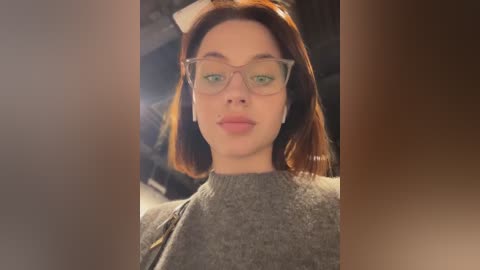 Media: A video of a young woman with light skin and straight, shoulder-length red hair, wearing glasses and a grey sweater. She stands indoors with a blurred background.