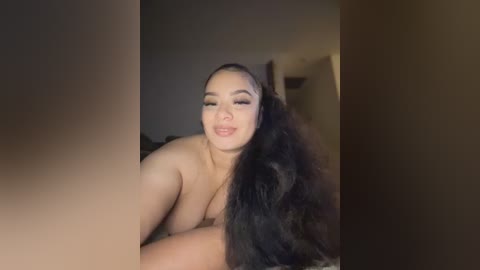 Media: Video of a young, light-skinned woman with long, dark hair, partially covered by a blanket, lying on a bed in a dimly lit room. She has a slight smile and is topless.