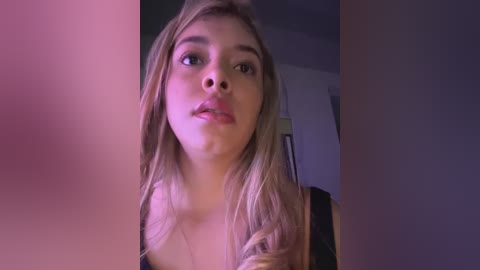 Media: Video of a young Caucasian woman with long, wavy blonde hair, light skin, and a neutral expression, wearing a black top, indoors with dim lighting.