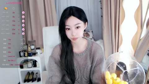 Media: Video of a young East Asian woman with long black hair, wearing a sheer grey sweater, seated indoors by a window with a bowl of yellow candies.