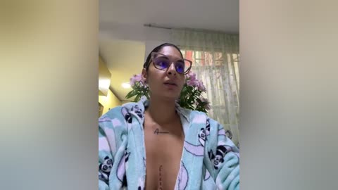 Media: A video of a young woman with medium skin tone, wearing sunglasses and a light blue robe with panda pattern, standing in a softly lit room with white curtains.