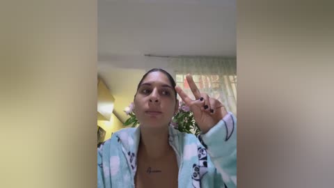 Media: Video of a young woman with medium skin tone and dark hair tied back, wearing a light blue and white striped robe. She makes a peace sign with her right hand, standing in a bright, softly lit room with green curtains.