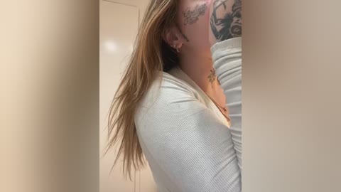 Media: A video of a young woman with long, straight blonde hair, wearing a white ribbed top, and visible tattoos on her neck and shoulder, standing in a beige room.