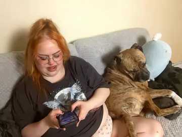 Media: A video of a plus-sized woman with red hair and glasses, wearing a black T-shirt, playing a video game on a couch with a brown dog and a blue stuffed animal.