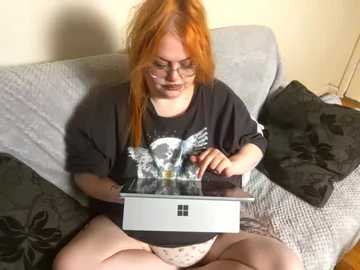 Media: Video of a pale-skinned, red-haired woman with glasses, wearing a black T-shirt, and light-colored panties, sitting on a gray bed with dark pillows, using a Microsoft Surface Pro tablet.