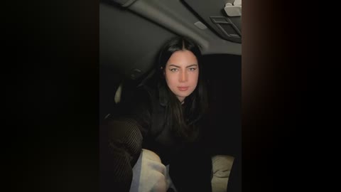 Media: Video of a woman with long black hair, wearing a black sweater, seated in a dark car, looking pensive, with dim lighting.