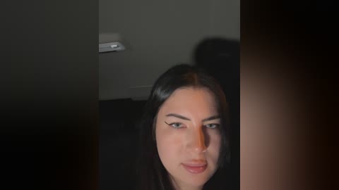 Media: Video of a young woman with long black hair and striking blue eyes, partially obscured by a dark object. The background is dimly lit with a white wall and a small white light fixture.