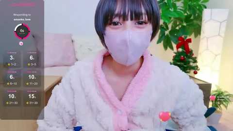 Media: Video of an Asian woman in a pink, fluffy robe, wearing a white face mask, standing indoors with a Christmas tree in the background.