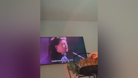 Media: A video of a young girl with light skin and dark hair watching a television screen displaying a musical performance. A hand holds a violin, and a potted plant sits on a black metal chair in the background.