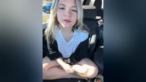 Media: Video of a young Caucasian woman with light blonde hair, wearing a black jacket and white shirt, sitting in a car, looking thoughtful.