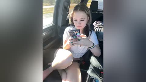 Media: A candid video of a young woman with fair skin, brown hair, and medium build, sitting in a car, taking a selfie with a smartphone. She wears a white top and shorts. The car interior and road outside are visible.