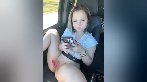 Media: Video of a young woman with light skin, short blonde hair, and green nail polish, sitting in a car, holding a smartphone, wearing a white shirt and no pants, exposing her shaved vulva.