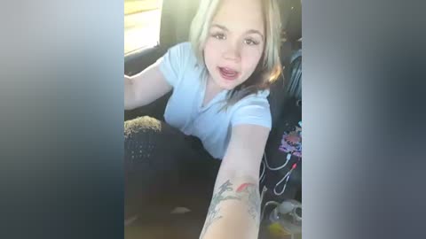 Media: Video of a young Caucasian woman with shoulder-length blonde hair, wearing a white t-shirt and a black knit sweater. She has a tattoo on her forearm. The car interior is visible with sunlight streaming in.