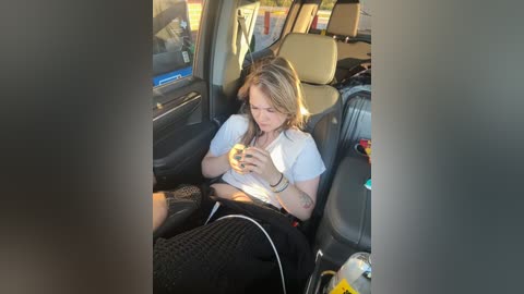 Media: Video of a blonde woman with medium build, wearing a white shirt and black pants, drinking from a bottle while seated in a car, surrounded by clutter and a blurry window view.