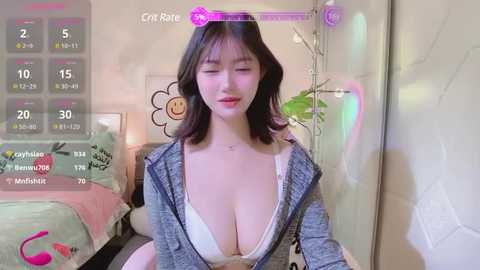 Media: Video of a young East Asian woman with fair skin, long black hair, and a large bust, wearing a partially unbuttoned gray shirt, sitting in a pink room with a bed, a plant, and a game screen overlay.