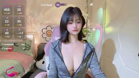 Media: Video of an Asian woman with straight black hair and fair skin, wearing a grey hoodie unzipped to reveal cleavage, seated in a pastel-colored room with a bed and plush toys.