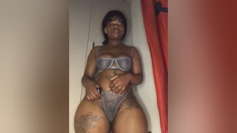 Media: Video of a dark-skinned woman in sheer, gray lingerie, with a tattooed thigh, lying on a bed with red curtains, her eyes closed.