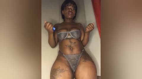 Media: Video of a dark-skinned woman with short hair, wearing sheer gray lingerie, sitting in a closet. She holds a blue object in each hand, with visible tattoos on her arms and thighs.