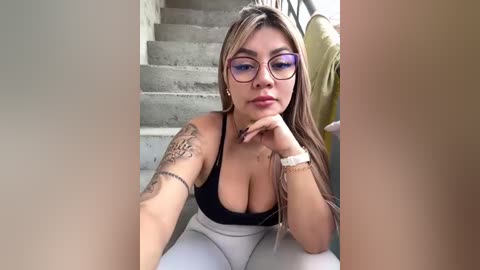 Media: Video of a light-skinned woman with long, straight blonde hair, wearing glasses and a black tank top, showcasing a tattoo on her left arm. She is sitting on stairs, looking at the camera.