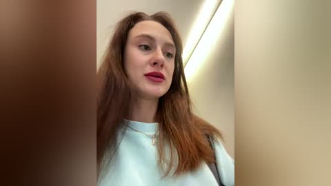 Media: A video of a young Caucasian woman with fair skin, long brown hair, and light blue eyes, wearing a light blue shirt. She stands in a brightly lit hallway with beige walls and fluorescent lighting.