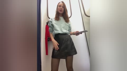 Media: A video of a woman in a light blue blouse and black skirt standing in a narrow airplane aisle, holding a smartphone.