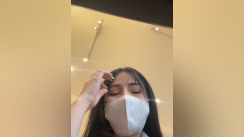 Media: Video of an Asian woman with long black hair, wearing a white face mask, adjusting her hair in a beige-walled bathroom with a black-framed mirror and ceiling lights.