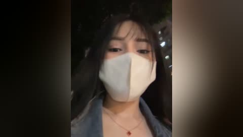 Media: A video of a young woman with long black hair, wearing a white face mask, a denim jacket, and a red necklace. The background is dark and blurred.