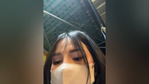 Media: Video of a young Asian woman with long black hair, wearing a white mask and a black top, looking down. The background shows a corrugated metal ceiling and blurred surroundings.