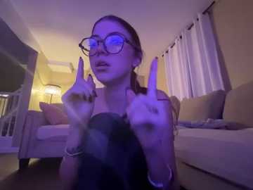 Media: Video of a young woman with light skin, glasses, and brown hair in a ponytail, sitting on a beige couch, flipping off the camera in a dimly lit living room with a white lamp and curtains.