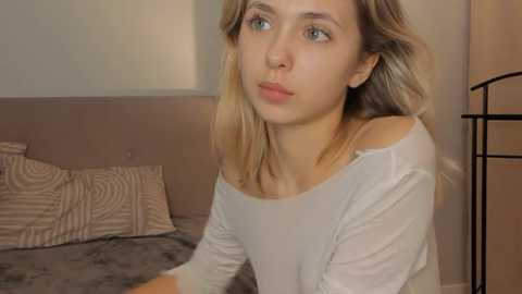 Media: Video of a blonde woman with blue eyes, wearing a white off-shoulder top, sitting on a beige couch with patterned pillows, looking contemplative.