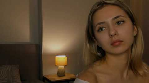 Media: Video of a blonde woman with fair skin, light makeup, and a white off-shoulder top, looking contemplative in a dimly lit, modern bedroom with a wooden nightstand and lamp.