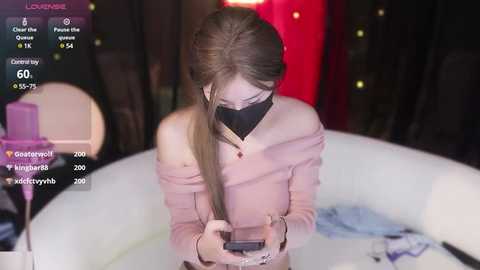 Media: Video of a young, fair-skinned woman with long, straight, light brown hair in a ponytail, wearing a pink sweater and black mask, sitting on a white chair, looking at her phone. Background includes dim lighting and blurred objects.