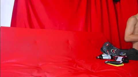 Media: Video of a nude person with light skin tone seated on a bright red couch, against a backdrop of red curtains. On the floor, a black handbag, smartphone, and a camera are visible.