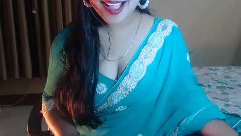 Media: A video of a smiling, South Asian woman with long, dark hair wearing a teal sari with intricate white embroidery, revealing a low neckline, sitting on a bed with floral sheets.