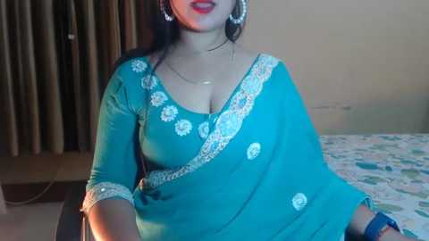 Media: Video of a woman in a teal sari with intricate white floral embroidery, wearing gold jewelry, red lipstick, and sitting indoors with beige walls and floral bedspread.