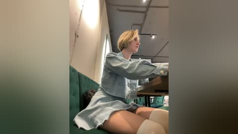 Media: Video of a blonde woman in a light blue hospital gown and white thigh-high stockings, sitting on a teal couch in a modern, well-lit room with exposed ceiling beams.