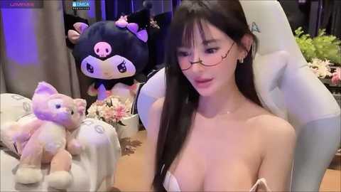 Media: Video of an East Asian woman with long black hair, glasses, and bare shoulders, seated in a gaming chair. Background features plush toys and a green plant.