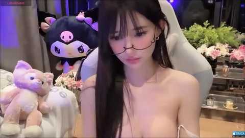 Media: Video of a young East Asian woman with long black hair, wearing glasses, and topless, sitting in a room with plush toys, flowers, and a dark background.