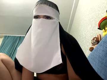 Media: Video of a person wearing a white veil with black trim, covering their face and shoulders, against a light blue textured wall, with a blue curtain in the background.