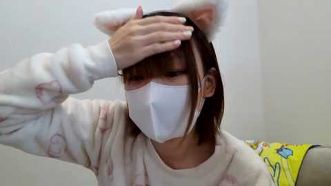 Media: Video of an Asian woman in a white pajama top with cat ears, wearing a face mask, and covering her head with her hand, in a plain room with a yellow blanket and a white couch.