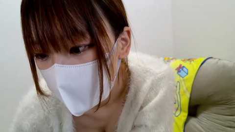 Media: A video of a woman with light skin and brown hair, wearing a white surgical mask, beige fur coat, and yellow floral-patterned blouse, indoors against a plain white background.