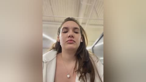 Media: Video of a young woman with light skin, brown hair, and a neutral expression, wearing a white jacket and a pendant necklace, standing in an indoor setting with a white ceiling and fluorescent lighting.