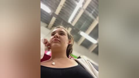 Media: Video of a young woman with light skin and brown hair, wearing a black top, looking up with a neutral expression. Background features a gymnasium with high ceiling and fluorescent lighting.