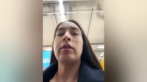 Media: Video of a woman with long dark hair, wearing a black jacket, taken from a low angle, indoors under fluorescent lights.