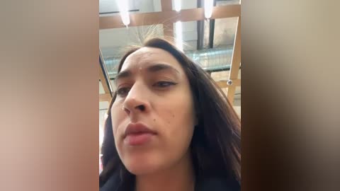 Media: Video of a young woman with olive skin, long dark hair, and a small mole on her right cheek, taken in a modern, well-lit indoor space with wooden accents and glass partitions.