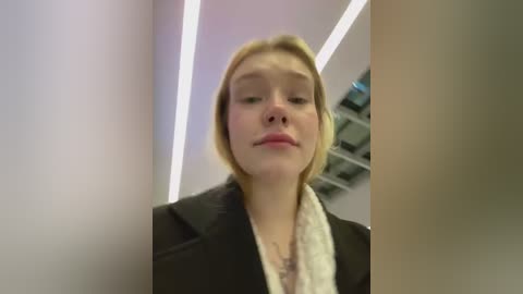 Media: Video of a young woman with light skin and short blonde hair, wearing a black jacket, standing in a modern, white-walled corridor with fluorescent lighting.