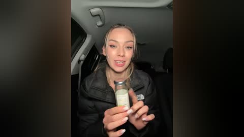 Media: Video of a blonde woman in a black jacket holding a glass jar with a white substance inside, seated in the backseat of a car.