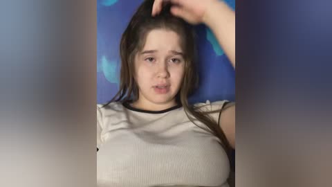 Media: Video of a young woman with long brown hair, wearing a white ribbed top, sitting against a blue background, looking tired and holding her hair.